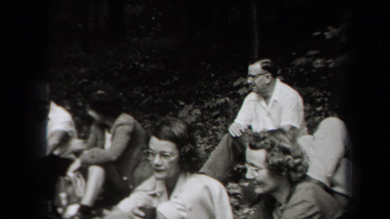 Adults sitting on the forest floor during a picnic, with trees and foliage behind them.