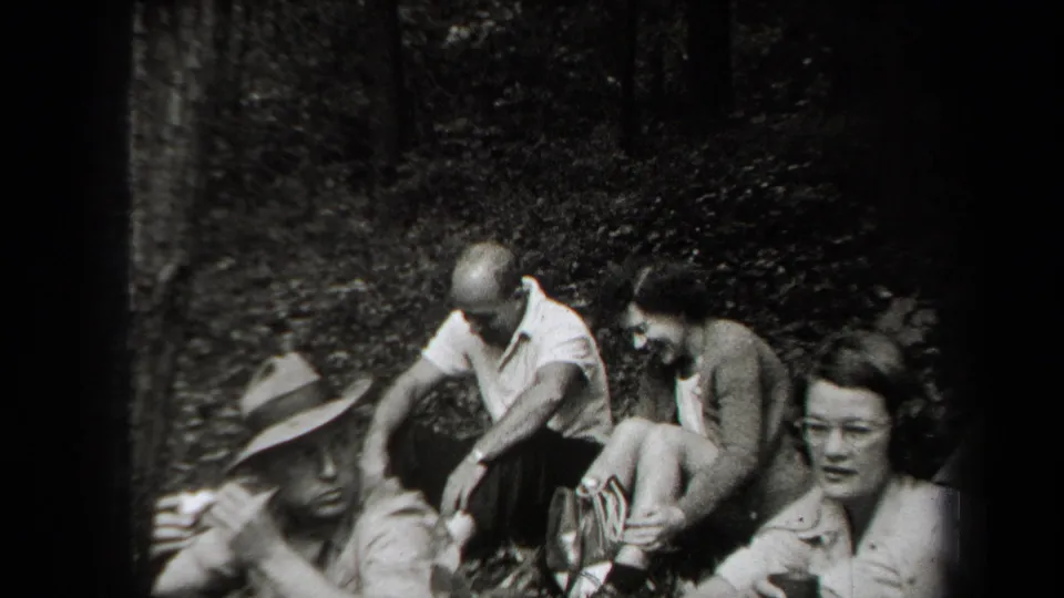 1938 New York home-movie frame showing adults resting in a wooded picnic area, with a brimmed hat and cups visible.
