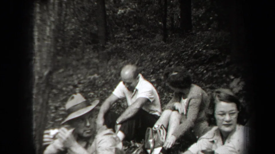 1938 New York home-movie frame of adults sitting close together on the forest floor during an informal outdoor meal.