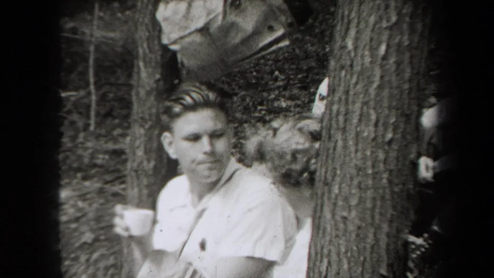 1938 New York home-movie frame with a man holding a cup near a tree as other adults sit in the wooded picnic area.