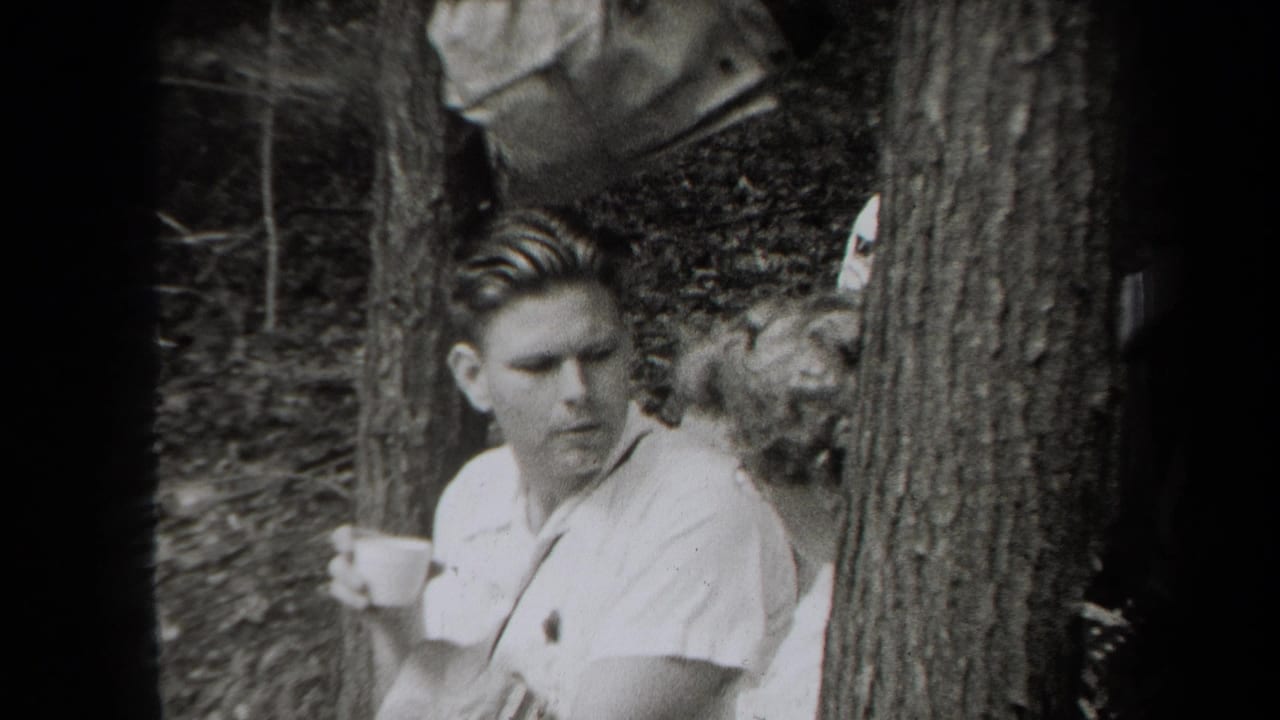 1938 New York home-movie frame of a young man in a light shirt holding a cup among trees, with another person partly obscured behind him.