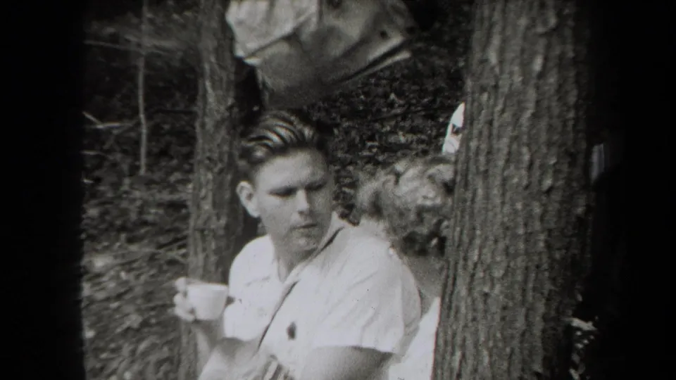 1938 New York home-movie frame of a young man in a light shirt holding a cup among trees, with another person partly obscured behind him.