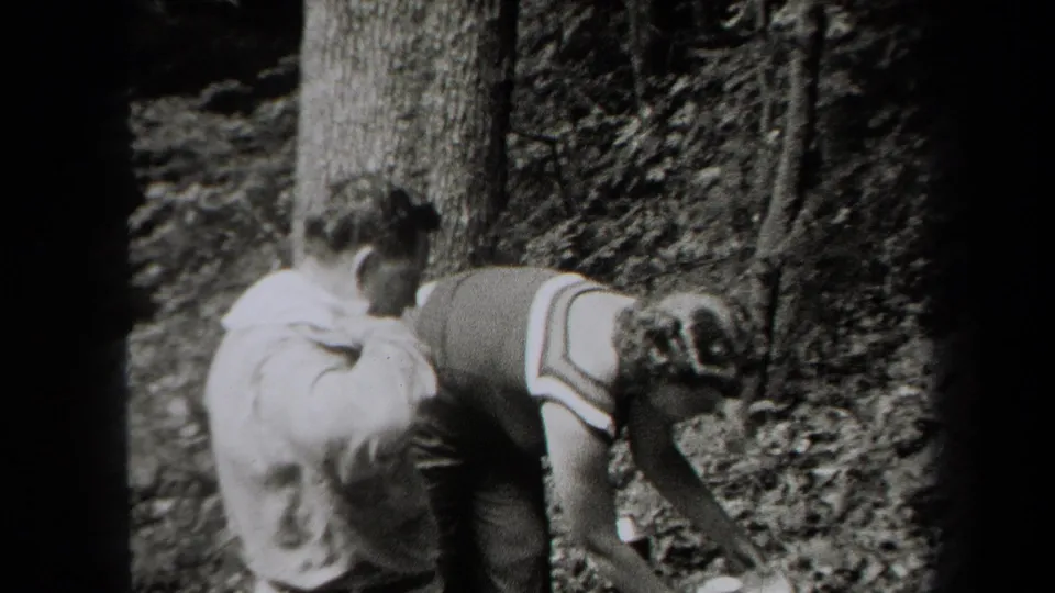 1938 New York 8mm frame showing two young men bending beside trees, with cups visible near the ground during an outdoor outing.