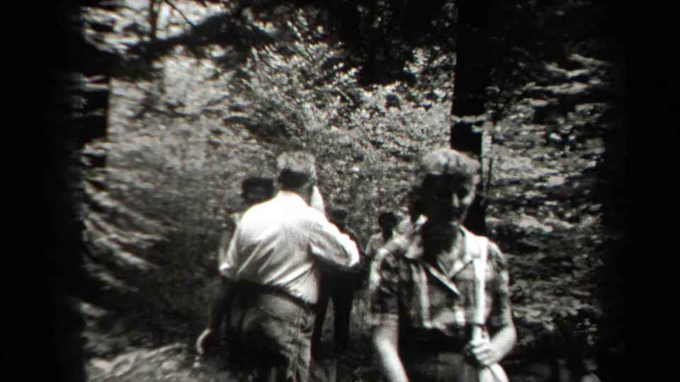 Black-and-white frame showing adults moving past the camera on a shaded New York woodland path, with foliage and tree shadows behind them.