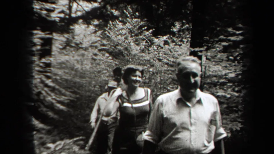 Two adults, including an older man in a light shirt and a woman in a dark dress, walk toward the camera on a wooded New York path in 1938 footage.