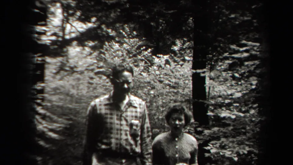 Two adult hikers in 1938 New York stand near a forest path, with dense leaves and tree trunks around them.