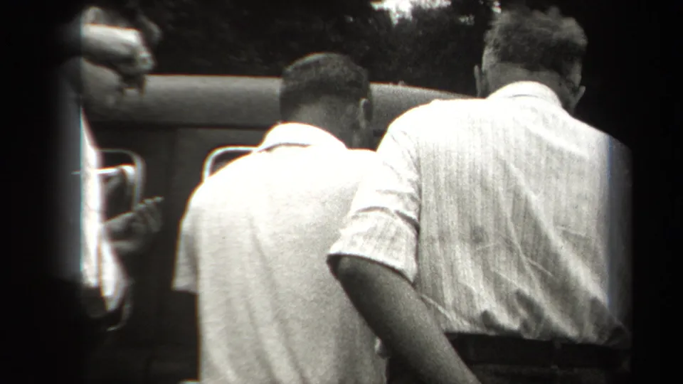 In 1938 New York home-movie footage, men gather beside a dark automobile, seen from behind in a tight black-and-white frame.