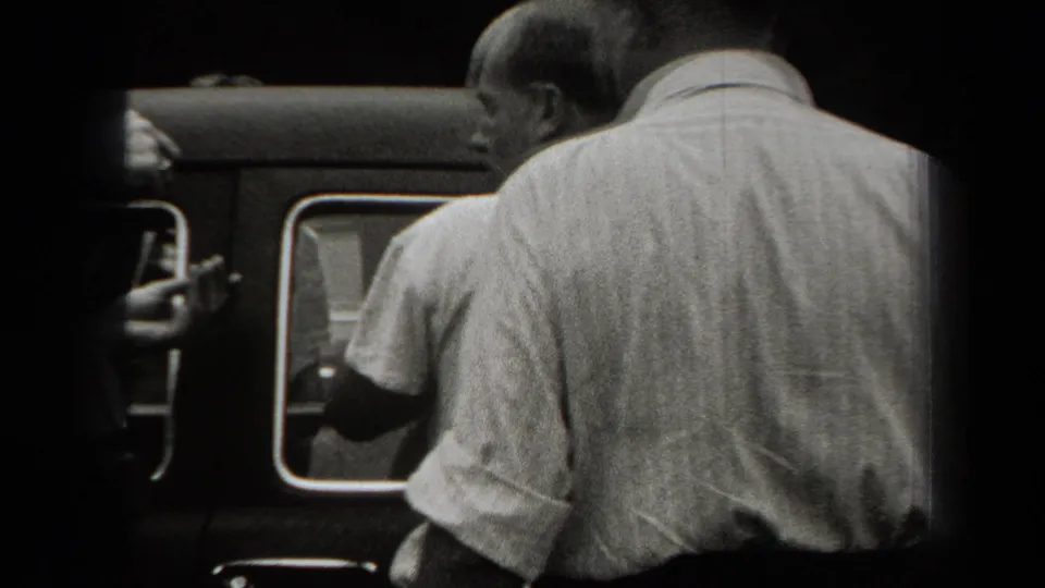 Two men stand close to the side of a dark automobile in supported 1938 New York footage, with a partial figure near the window.