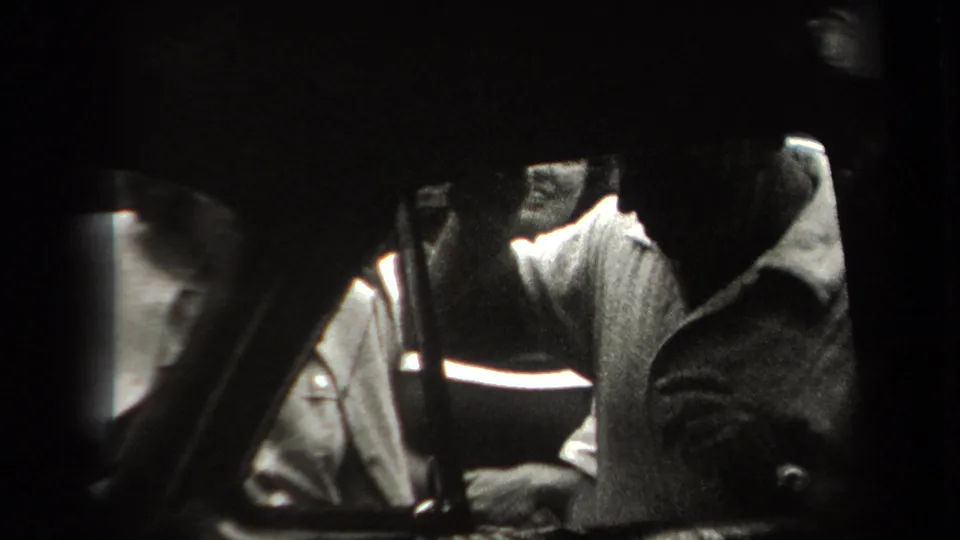 A tightly framed 8mm view shows a man bent near the automobile, with another person partly seen at the edge of the shot.