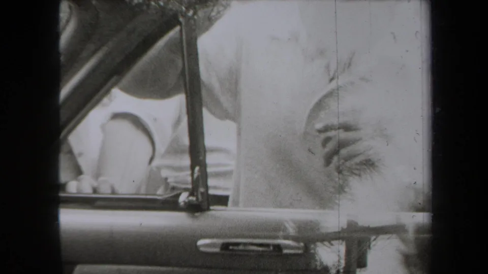 A man in a light shirt stands beside the automobile window in 1938 New York footage, while nearby figures are partly cropped by the frame.