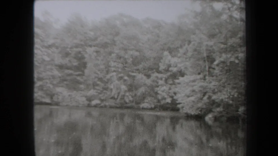 Black-and-white 1938 New York home-movie frame of a still lake bordered by trees, rocks, and a wooded slope.