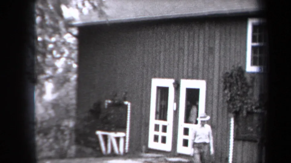 In 1938 New York home-movie footage, a man in a brimmed hat stands outside a clapboard farmhouse with white double doors and plants nearby.