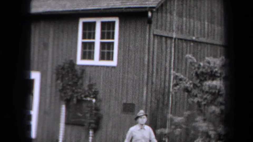 Black-and-white 8mm frame from New York in 1938 showing the man partly framed below a farmhouse wall, window, shrub, and doorway edge.