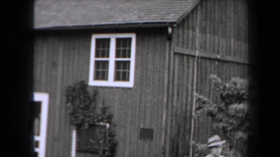 1938 New York home-movie frame showing a farmhouse exterior with a white-trimmed window, shrubs, and the man's hat and shoulders at the lower edge.