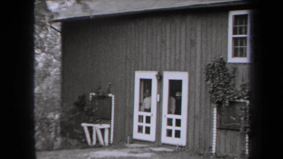 1938 New York 8mm frame showing a rustic wooden cabin exterior with white-trimmed doors, window, plants, and a small table.