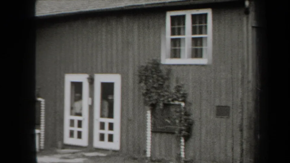 1938 New York home-movie frame of a wooden cabin entrance with white-framed doors, upper window, and foliage along the wall.