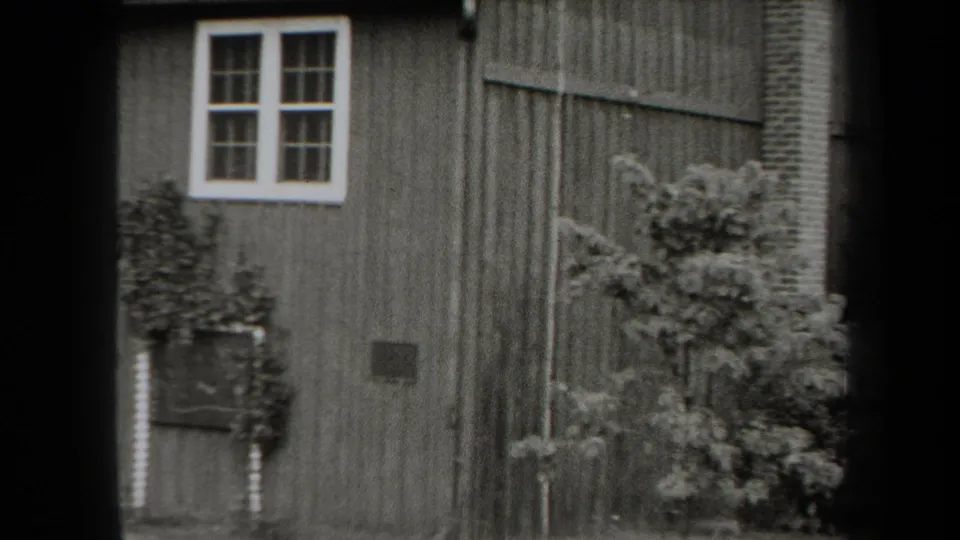 1938 New York 8mm frame showing the cabin side wall with a white-trimmed window, planter, vent, chimney, and dense shrubs.