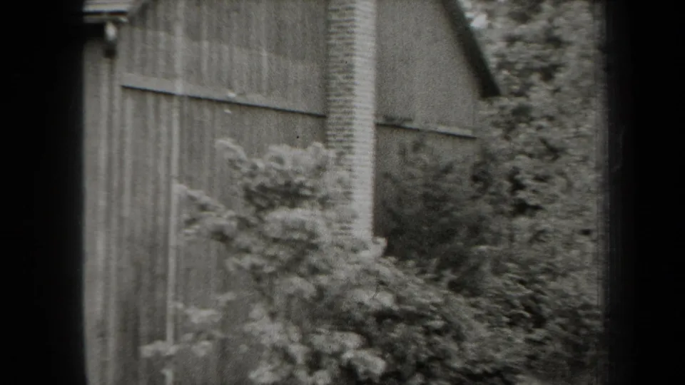 1938 New York home-movie frame with a cabin side wall, brick chimney, shrubs, roofline, and soft black-and-white 8mm texture.