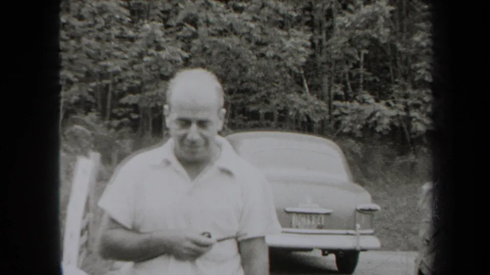 1938 New York home-movie frame with a man in a light polo shirt near a parked 1930s-style automobile in a wooded setting.