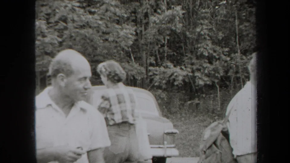 1938 New York home-movie frame showing adults around a parked car in a wooded roadside or picnic setting.