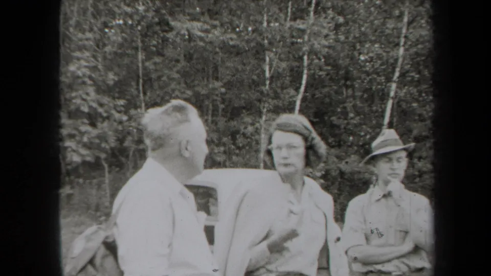 1938 New York home-movie frame of adults conversing near a car, with a woman facing a man and another man in a brimmed hat nearby.
