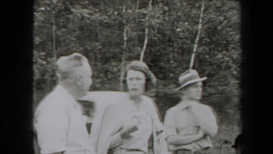1938 New York home-movie frame showing adults in period clothing talking beside a car at the edge of woods.