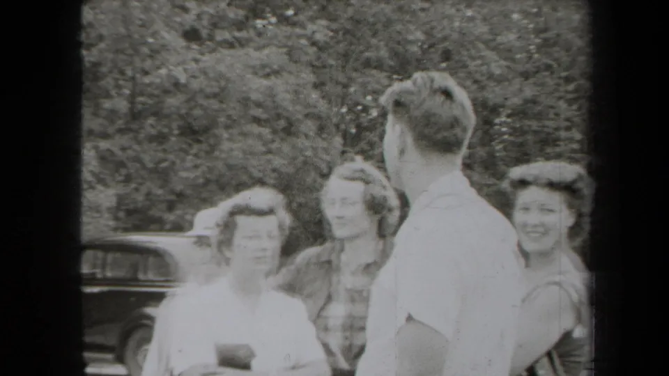 1938 New York frame of adults conversing beside a vintage automobile in a wooded outdoor setting.