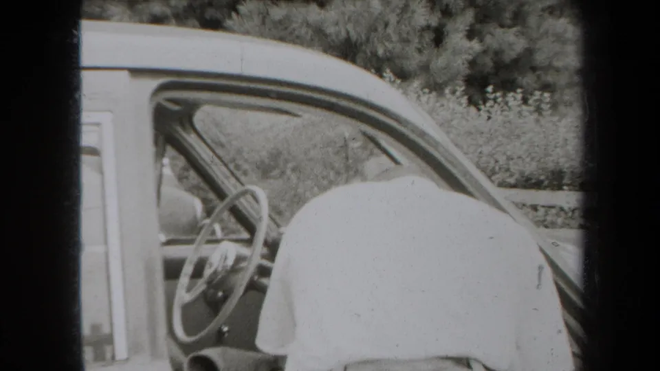 Black-and-white 8mm frame showing the side of a 1930s-era car with steering wheel and partial figure visible.