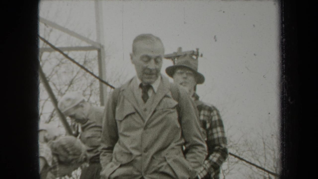 Two men outdoors among leafless trees, one in a suit and tie and one in a hat behind him.