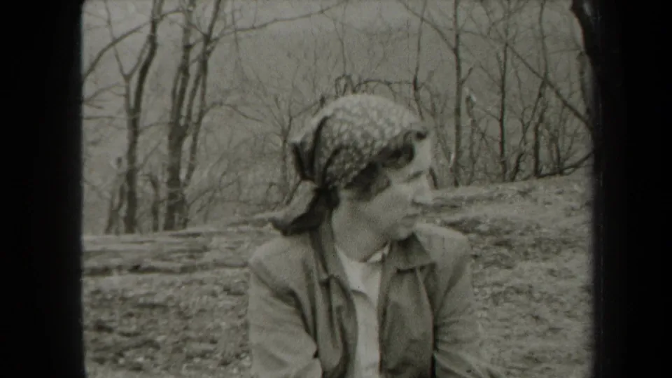 1938 New York home-movie frame showing an adult woman in a patterned headscarf looking to the side in a wooded area.