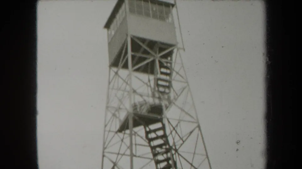 1938 New York home-movie frame showing a metal observation tower with stairs, filmed from below in a wooded outdoor setting.