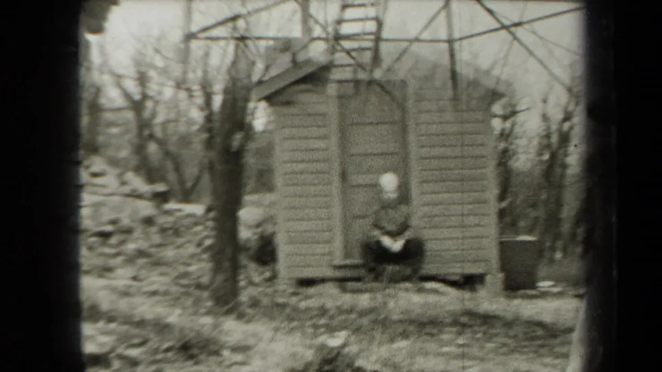 A small wooden shed in a leafless wooded setting, with a seated person in a light cap near the doorway, consistent with the 1938 New York metadata.