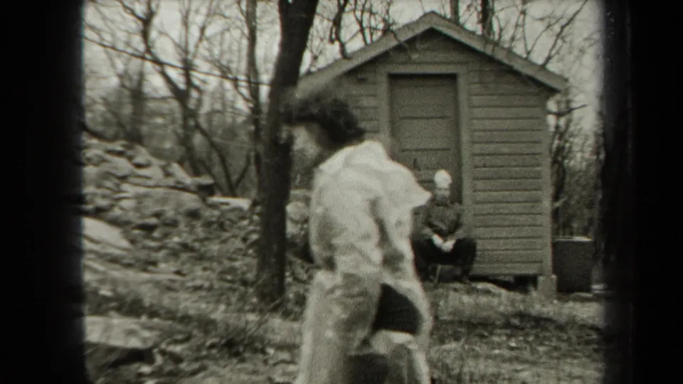 A person in a light coat crosses the foreground near a small shed, while another person sits by the doorway in wooded New York footage from 1938.