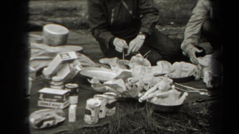 Outdoor poultry work with a seated person handling a chicken near pots, tins, boxes, and other selling or preparation supplies.