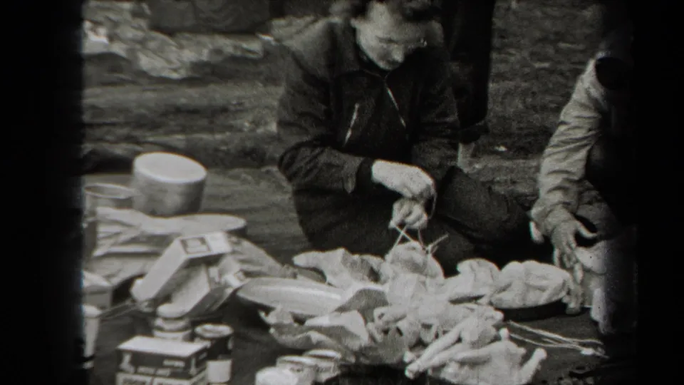 A man in dark clothing handles poultry on the ground, with cookware, boxes, and small containers visible around the work area.