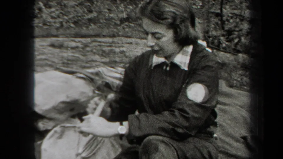 A woman in a dark jacket with a light sleeve patch prepares food on a blanket during a 1938 New York camping scene.