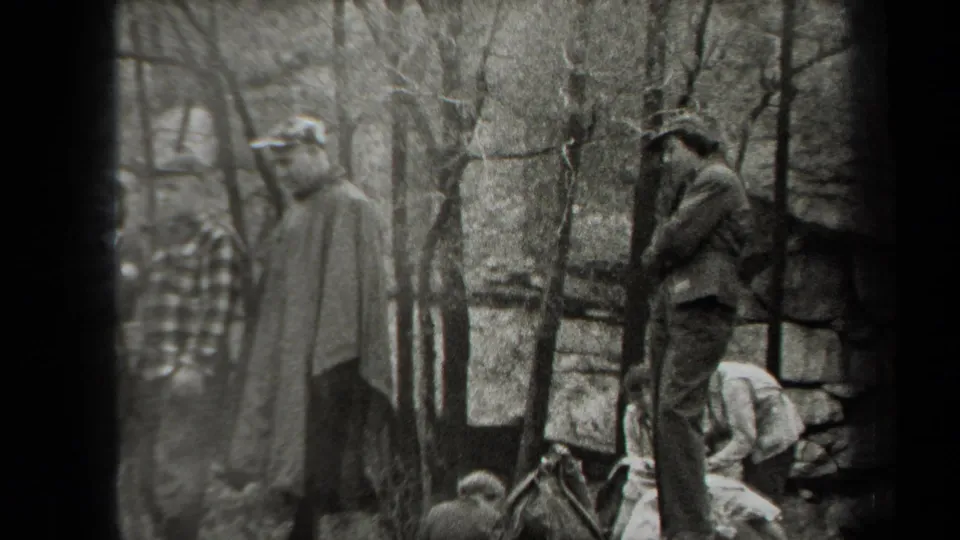 Several campers gather among trees, with men in hats and one person bending near the ground in 1938 New York home-movie footage.