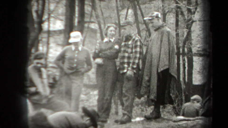 A small group of campers, including men in hats and a plaid shirt, stands and sits together in a wooded New York setting.