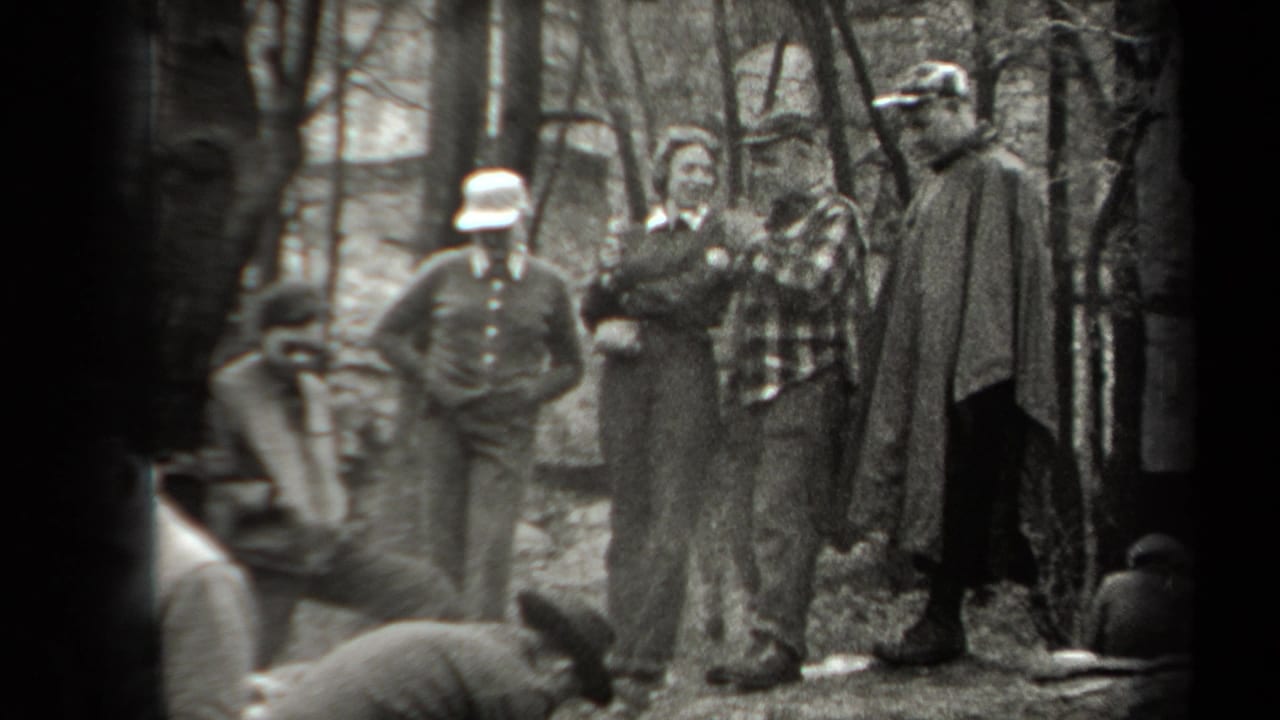 In 1938 New York home-movie footage, adults in hats and jackets stand in a wooded area near smoke from a small fire.