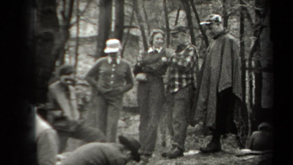 In 1938 New York home-movie footage, adults in hats and jackets stand in a wooded area near smoke from a small fire.