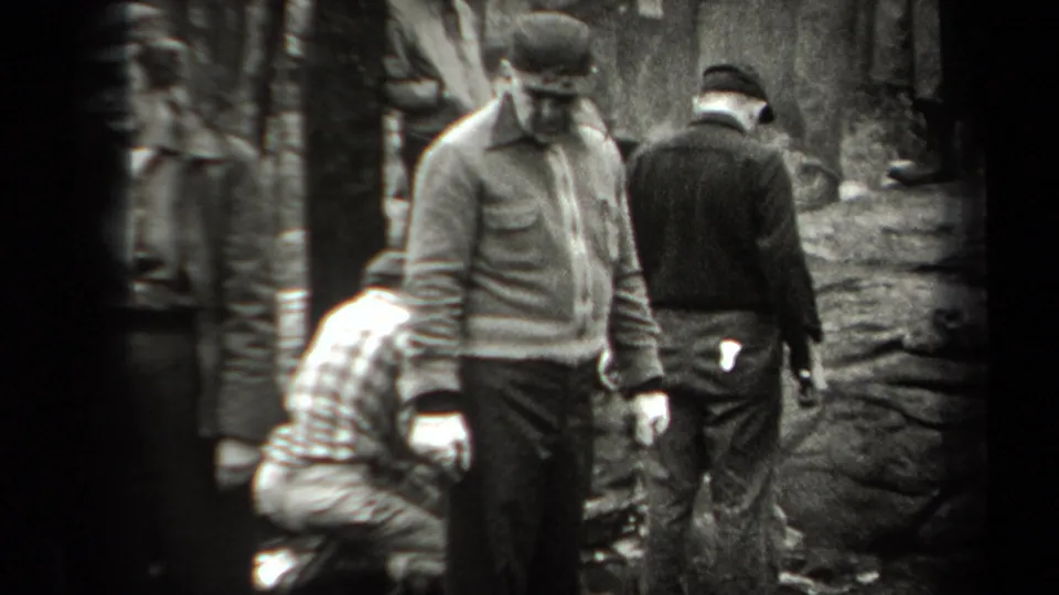 Adults in caps and jackets move through a wooded New York setting, with one figure partly bent near the ground.