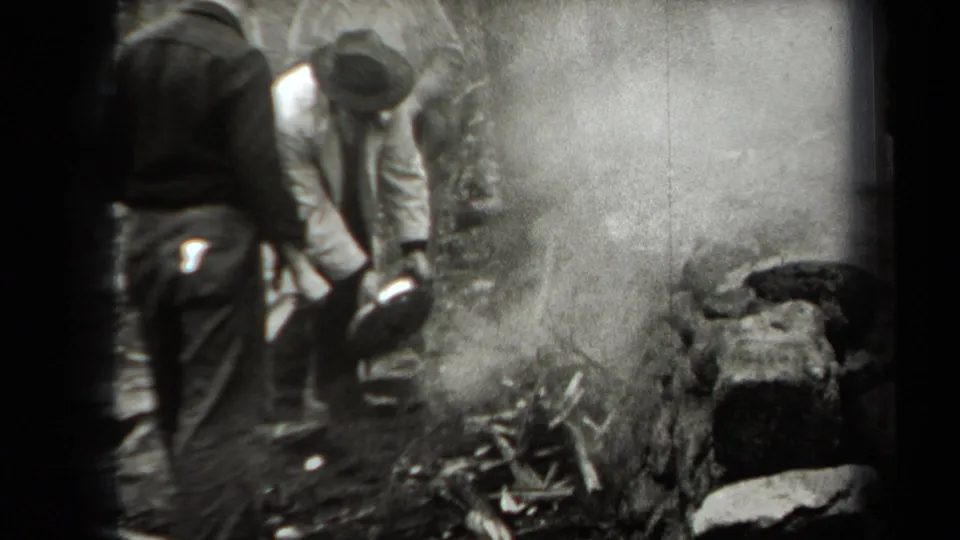 A man bends toward a smoky small fire in the wooded scene while another adult stands nearby in period clothing.