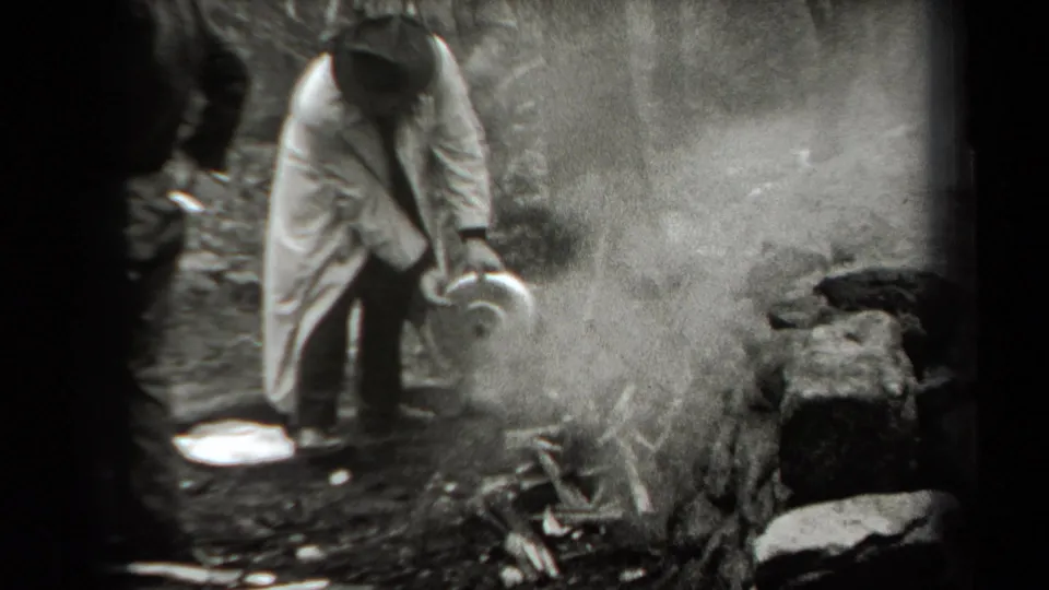 In the wooded 1938 New York scene, a man in a long coat appears to fan smoke rising from a small fire near rocks.