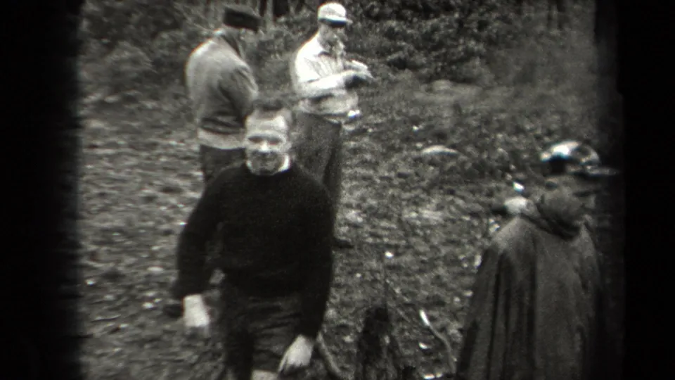 Men in coats and caps gather in a New York wooded setting in 1938, with one dark-sweatered man near the center foreground.