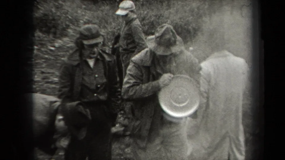 Men in a 1938 New York wooded home-movie scene gather around a cooking area as one figure holds a round metal pan.
