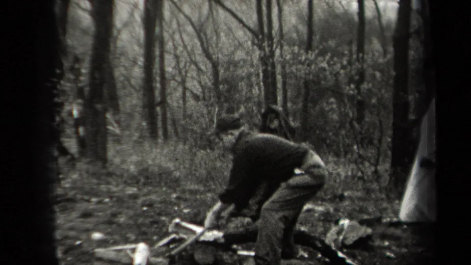 In 1938 New York home-movie footage, a man bends over kindling and logs in a wooded area as other men stand nearby.