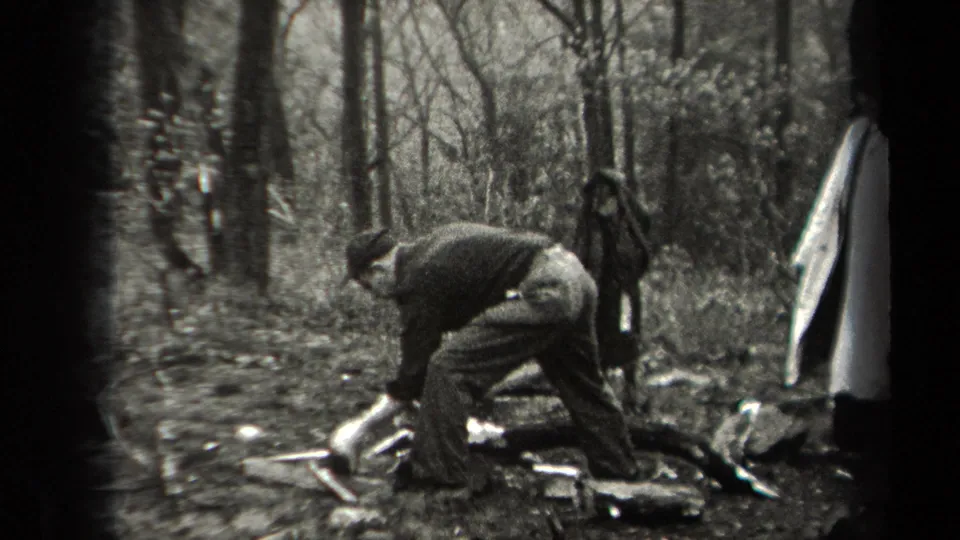 A man in period outdoor clothing leans over a pile of wood and debris in a wooded New York setting, with another figure standing behind.