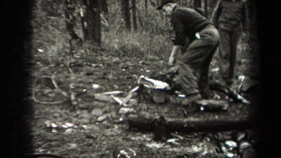 A man bends close to logs and kindling in the forest, with another person partly framed beside him in the 1938 home-movie scene.