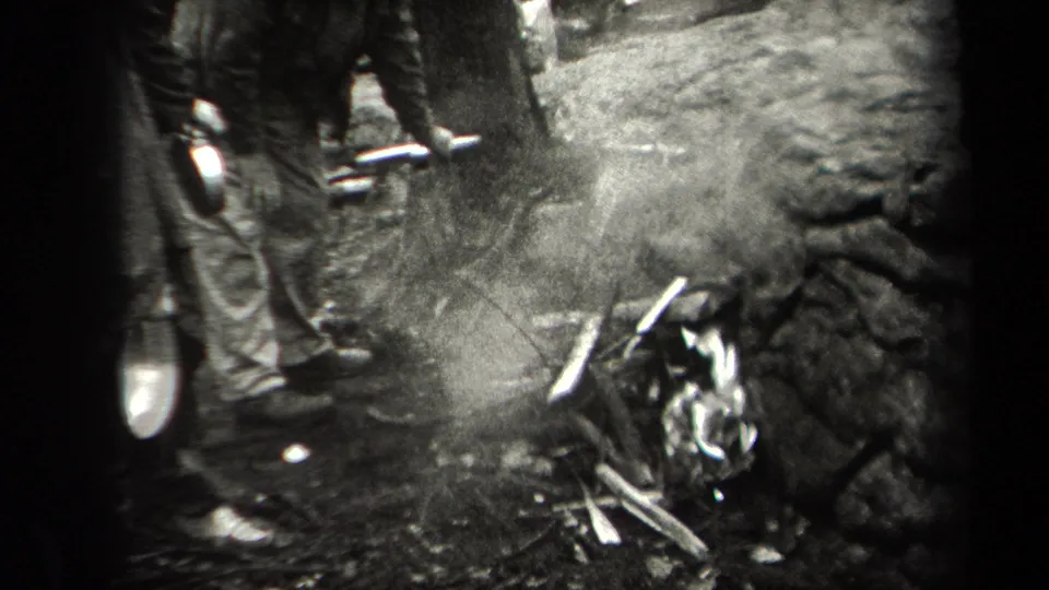 Smoke rises from the campfire area in the wooded New York scene as men stand nearby and logs lie along the forest floor.
