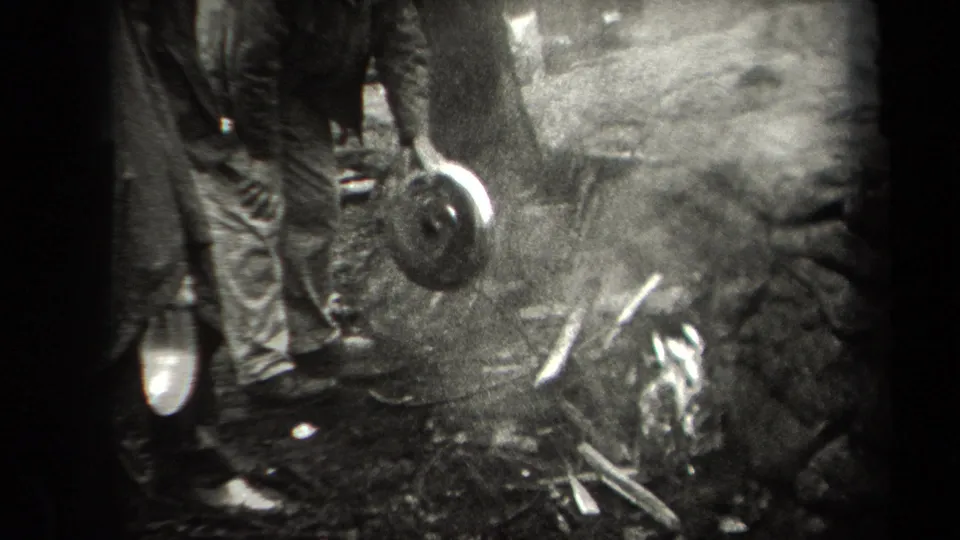 1938 New York 8mm frame showing partially seen adults beside a rough outdoor fire pit with a suspended chicken and cooking tools.